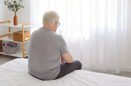 Backside view on a senior man sits thoughtfully on bed in the morning, greeting a new day in his bedroom. Reflective moment at home, quiet contemplation after of waking up to a new routine.の写真素材