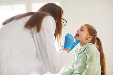Female doctor pediatrician laryngologist with tongue depressor asks little child girl to open mouth for throat, larynx and pharynx diagnostic examination for strep bacteria, flu, or pharyngitisの写真素材