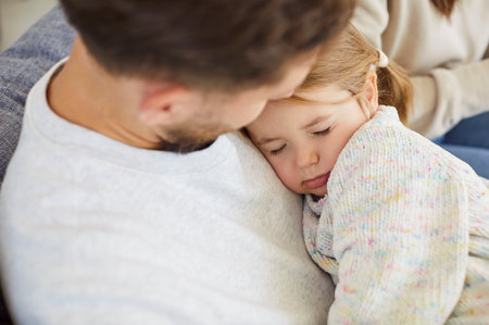 Loving and caring father cradles sleeping little daughter in hands while resting together on the comfortable sofa at home living room. Family affection, warmth, and peaceful moments.の写真素材