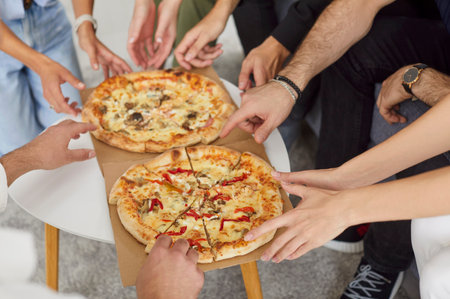 Close-up shot of friends hands grabbing pizza from the table during a party at home. The fun and casual atmosphere captures the group enjoying lunch or dinner together with connection.の写真素材