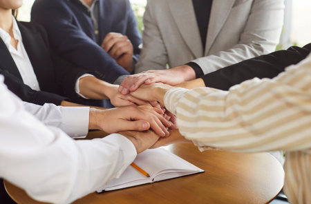 Business people group putting their arms together in a pile standing at the desk on workplace in office. Stack of hands of company employees. Unity, partnership and teamwork concept.の写真素材