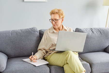Mature happy freelance business woman in glasses working at home on sofa, using laptop, resting on couch, typing on computer, smiling, laughing, looking at copybool, senior lady shopping online, relaxの写真素材