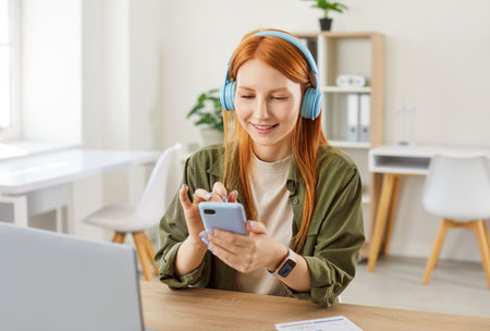 Portrait of smiling young redhead girl in casual clothes wearing headphones sitting at desk with laptop at home using mobile phone reading news in social media browsing app or working onlineの写真素材