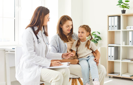 Little girl patient sitting on mother knee during visit to doctor in medical clinic. Friendly female pediatrician in lab coat talking to mother and child, providing support during health checkup.の写真素材