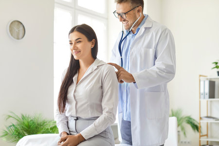 Doctor or nurse uses a stethoscope to listen to the breath and lungs of a patient during an exam in the clinic office. The medical professional provides attentive care during this important checkup.の写真素材