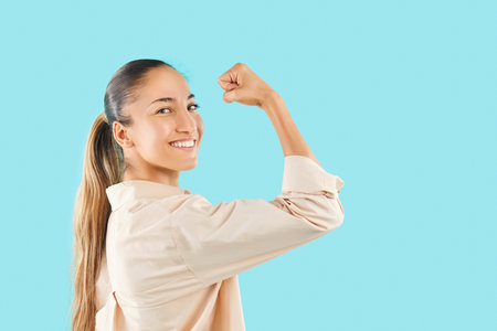 Portrait of a happy and positive woman showing strong bicep on a bent arm, looking at camera against a blue background. Her sporty and powerful pose highlights strength and enthusiasm.の写真素材