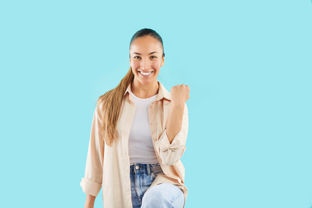 Portrait of an excited and happy woman celebrating success with a fist pump on a blue background. Her expression shows victory and luck, making a perfect symbol of positive achievement.の写真素材