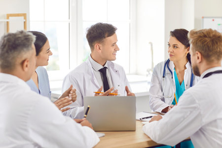Happy young team of confident doctors in white uniform sitting on workplace in hospital or clinic on meeting discussing diagnosis or planning treatment for patients. Medicine and health care concept.の写真素材