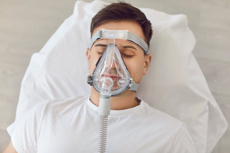 Man sleeping in bed at home, breathing through an oxygen mask. He receives medical treatment for asthma or sleep apnea, ensuring health and well being in a home healthcare setting.の写真素材