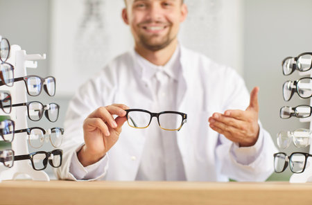 Portrait of young happy smiling male optician doctor holding eyeglasses in hands working in medical clinic sitting at desk on workplace. Ophthalmology, vision and eye health concept.の写真素材