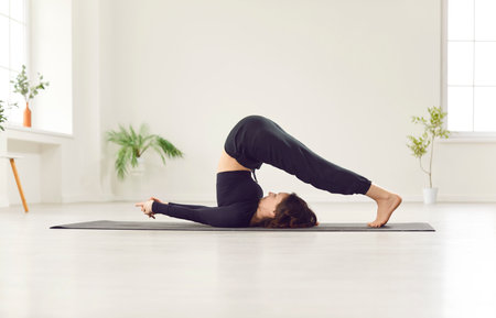 Sporty yogi woman wearing black sportswear practicing yoga, doing Halasana exercise, standing in Plough pose, beautiful girl working out at home sport or modern studio fitness for healthy lifestyleの写真素材