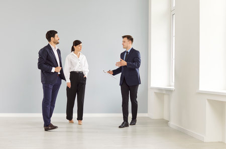 Male realtor showing new office, young family, happy couple in new empty room, real estate agent, clients at meeting, showing, selling buying property for rent, sale, discussing contract with customerの写真素材