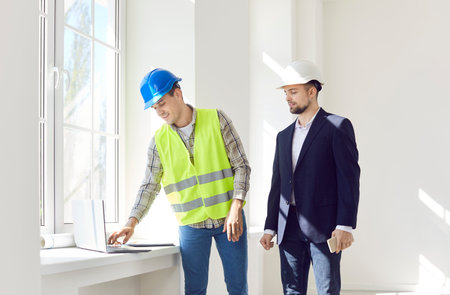 Two men in helmets look at a laptop at a construction site, close-up. Solving questions on engineering structures. House renovation concept. Coordination of work with the architectの写真素材