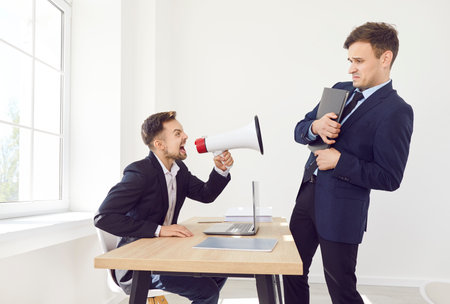 Angry boss sitting at the desk on his workplace holding megaphone and shouting at his young stressed employee standing in office. Businessman yelling at male worker. Dismissal concept.の写真素材