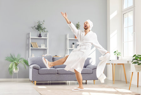 Portrait of happy funny excited young man in white bathrobe holding towel and having fun at home enjoying beauty spa day preparing for cosmetic procedures. Male skin and body care concept.の写真素材