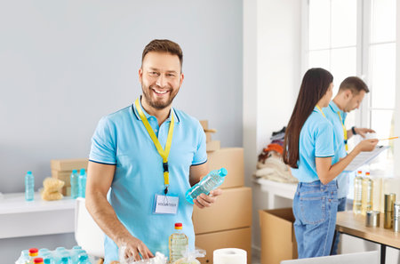 Portrait of young smiling man volunteer sorting donations of foodstuffs for charity food drive volunteering in food bank in charitable foundation helping poor people in need. Humanitarian aid conceptの写真素材
