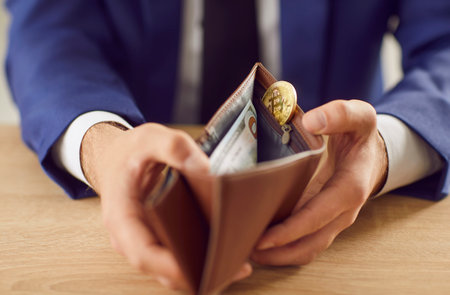Man opens up his wallet. Close up male hands holding leather wallet with paper dollar banknote and symbolic golden bitcoin inside on wooden table. Bitcoin, business, money, finance conceptの写真素材