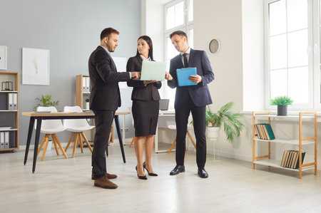 Team of three business people in suits standing in office interior, looking at laptop computer, and discussing web marketing project that they are working on togetherの写真素材