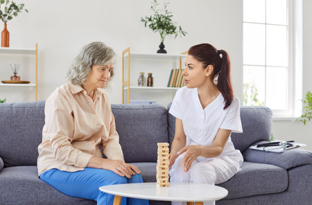 Senior patient plays board game together with nurse as dementia treating or preventing activity. Sick old mature woman with Alzheimers sitting on sofa with nurse, looking at wooden tower and thinkingの写真素材