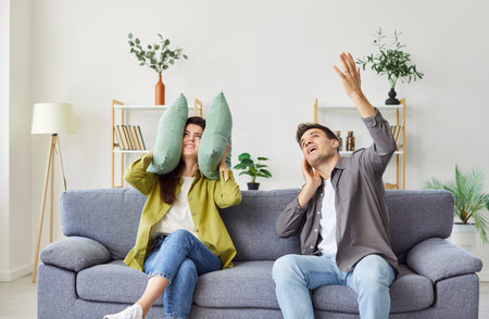 Annoyed young woman covering ears with pillow at home, blocking sound of man singing, speaking loud, suffering from noise, irritated girl hiding from loud boyfriend, noisy funny roommate neighborの写真素材