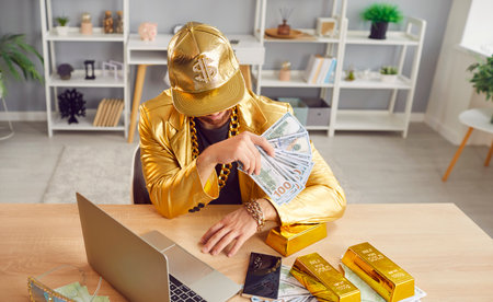 Top view of funny man sitting at the desk, holding dollars banknotes, working online using laptop. Rich guy in golden suit with money cash. Investment and business success concept.の写真素材