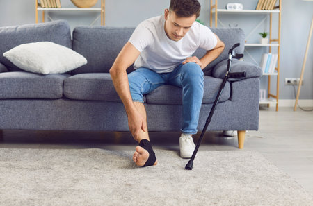 Unhealthy young man with foot trauma or sprain sitting on sofa next to the crutch, touching injured ankle with black elastic bandage and feeling pain. Man with injuries is being rehabilitated at home.の写真素材