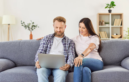 Family couple relaxes on the couch at home, using a laptop for online entertainment. They enjoy leisure time together, highlighting the internet, relaxation, and shared moments in a home environment.の写真素材