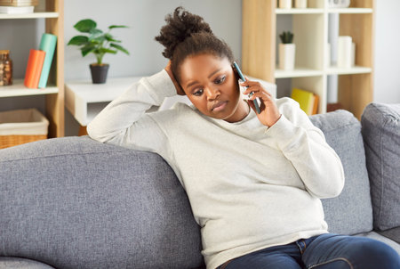 Portrait of a young african american girl talking by phone while sitting on sofa at home with upset worried face expression. Sad woman resting on couch with smartphone in living room.の写真素材