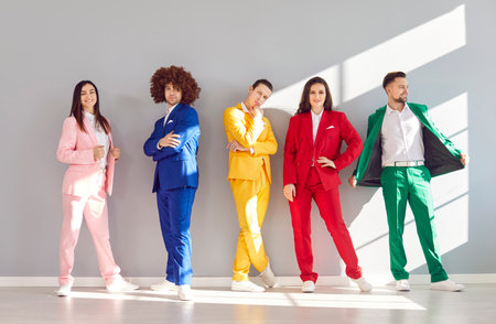 Group of funny cheerful people men and women in colorful bright stylish suits standing in a row on gray wall background looking at camera. Portrait of young friends in party fashion costumes.の写真素材