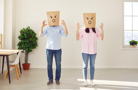 Family couple having a fight. Young man and woman having a conflict. Two people wearing paper bags with angry face expressions standing in the living room at home, quarreling and gesticulatingの写真素材