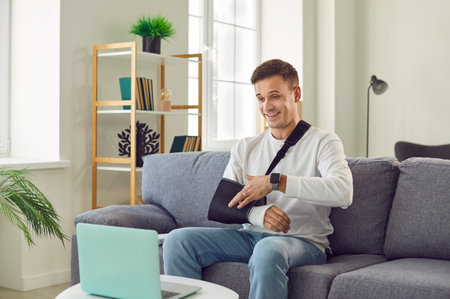 Happy young man with arm in a cast sitting on the couch at home and communicating on a laptop. Man in rehab after injury at home chatting on computer and smiling having online video call.の写真素材