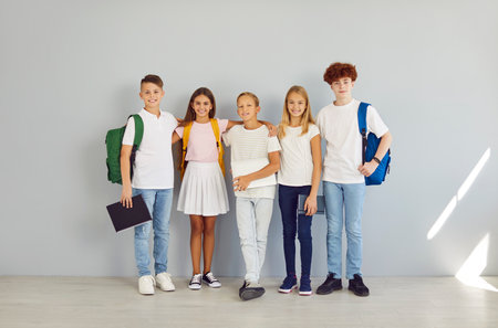 Smiling happy school students children looking cheerful at camera and hugging isolated on gray wall background. Boys and girls with backpacks standing in a row. Back to school concept.の写真素材
