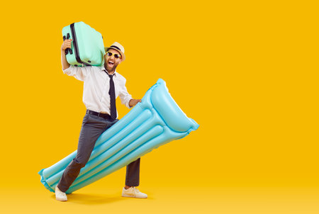 Happy business man traveling on summer vacation. Funny crazy man in office shirt, tie, sun hat and glasses carrying holiday green suitcase and blue rubber water mattress on yellow studio backgroundの写真素材