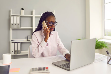 Young businesswoman at work. Beautiful African American business woman in glasses sitting at office table, talking on cellular phone and looking at laptop computer screen with serious face expressionの写真素材