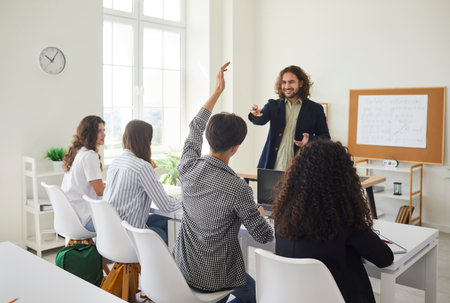 Group of high school students having class. Teenage boy sitting at a desk together with girls in the classroom and raising his hand up to ask the teacher a question. Back to school conceptの写真素材