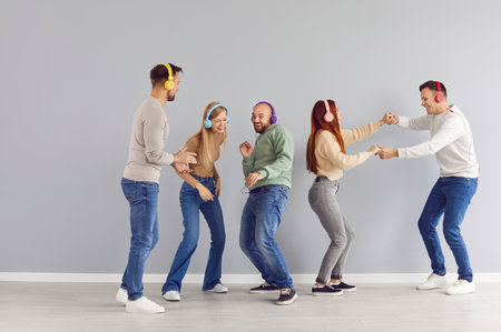 Happy friends enjoying party, listening to music, dancing and having good time together. Group of young people in colorful stereo headphones dancing by grey wall to playlist of favorite disco songsの写真素材