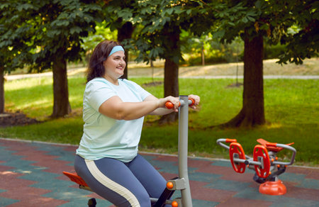 Happy beautiful smiling adult woman in sportswear having workout on sports ground in park and doing fitness exercises on red grey metal equipment, with green trees in backgroundの写真素材