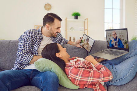 Joyful young married couple uses various gadgets to call their elderly parents online. Smiling Caucasian woman and man at home on sofa using laptop and digital tablet for online chat with family.の写真素材