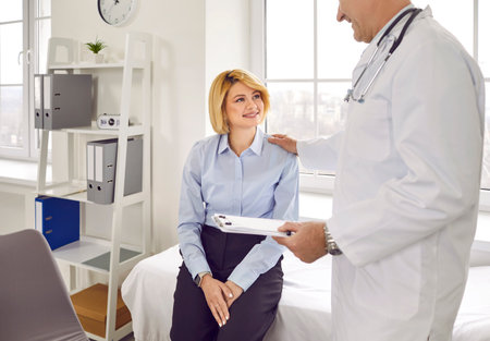 Woman patient having medical checkup at clinic. Happy young girl sitting on examination couch listening to friendly, supportive doctor in white coat telling about treatment and good recovery prognosisの写真素材