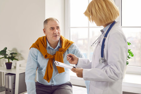 Mature man patient in medical clinic talking with female adult friendly doctor wearing stethoscope and holding report file with appointment and giving consultation to a man during medical examinationの写真素材