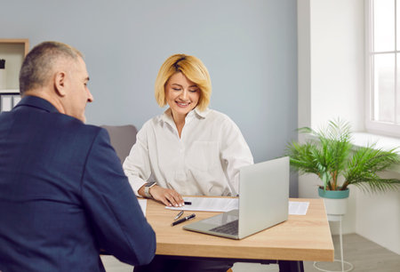 Professional loan broker or financial advisor meeting with client. Happy beautiful young woman sitting at office desk with senior man and showing him business offers on laptop computerの写真素材