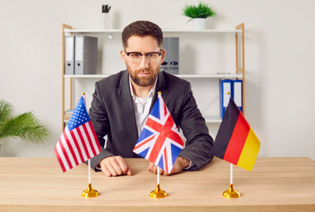 Pensive young man sitting at desk in office looking at flags of different countries. Businessman choosing foreign language or country to work or study in. Education or emigration conceptの写真素材