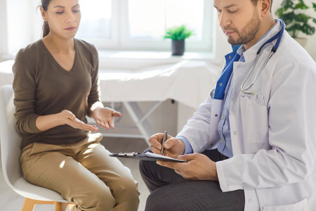 Doctor and patient during a medical appointment. Serious man physician in a white coat sitting on a chair, interviewing a woman patient and writing down her symptomsの写真素材