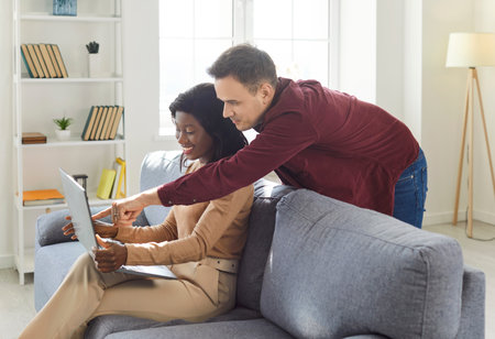 Multiracial young couple, interracial family at home using laptop online, pretty african american woman and caucasian man boyfriend reading message, working together at computer, choosing, orderingの写真素材