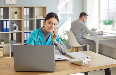 Friendly female nurse receptionist answering telephone call, sitting at desk with laptop computer, scheduling appointments, writing down notes in notebook, posing in medical clinicの写真素材