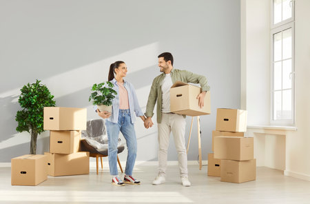 Happy joyful young couple standing with a lot of unpacking cardboard boxes in a new apartment holding hands and smiling celebrating moving day. Relocating, real estate, mortgage concept.の写真素材
