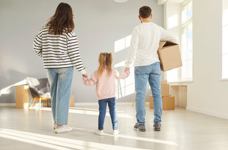 Young family enters their newly purchased home, full of excitement and anticipation. Rear view of parents holding their child hands and walking together to living room carrying cardboard box.の写真素材