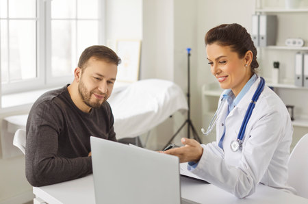 Female doctor in white medical uniform at laptop showing results or symptoms with male patient, GP or physician consulting client, gives recommendation at meeting in hospital, helping, explainingの写真素材