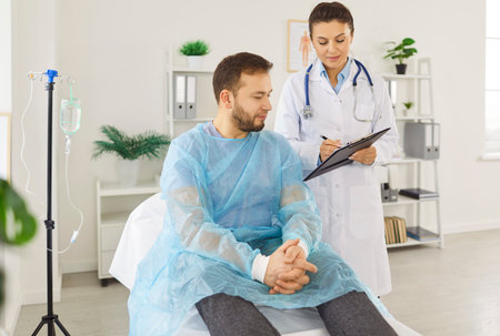 Female doctor consulting male patient in clinic chair, sharing good lab results, checking paper document, visiting clinic room for medical care, getting health advice, discussing treatment planの写真素材