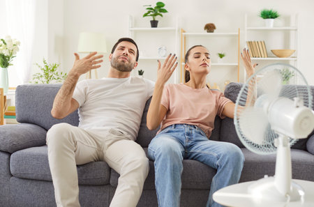 Portrait of young couple in casual clothes sitting on sofa in living room at home and suffering from summer heat in front of fan. Man and woman enjoying electric fan during heatwave.の写真素材